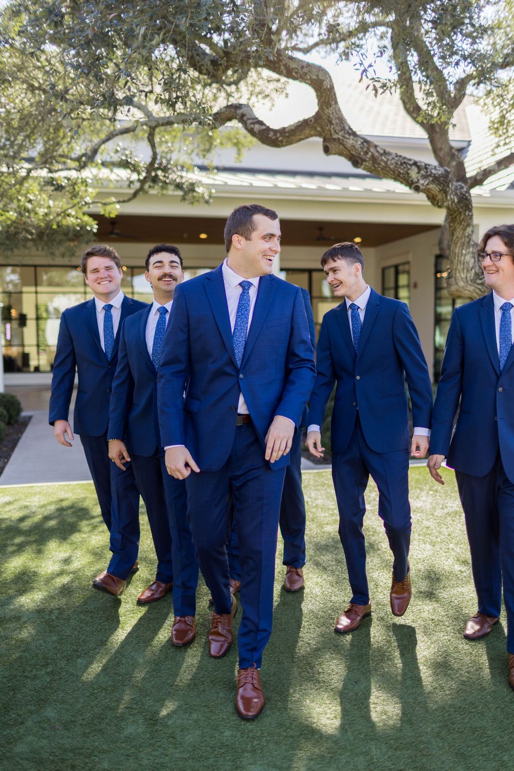 Five groomsmen in navy blue suits and brown dress shoes walk outdoors on green grass near a building.
