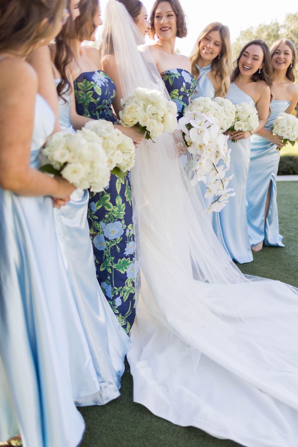 Bride in white gown with veil stands among bridesmaids in light blue and floral dresses holding white bouquets outdoors.