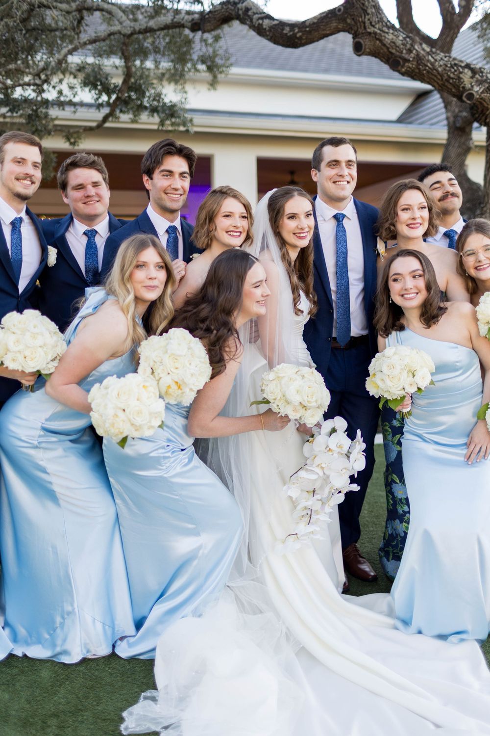 Bride in white gown with bridesmaids in light blue satin dresses and groomsmen in navy suits holding white bouquets.
