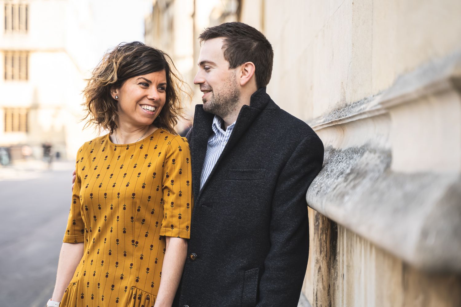 A smiling couple in stylish autumn outfits share a joyful moment leaning against a stone wall outdoors.