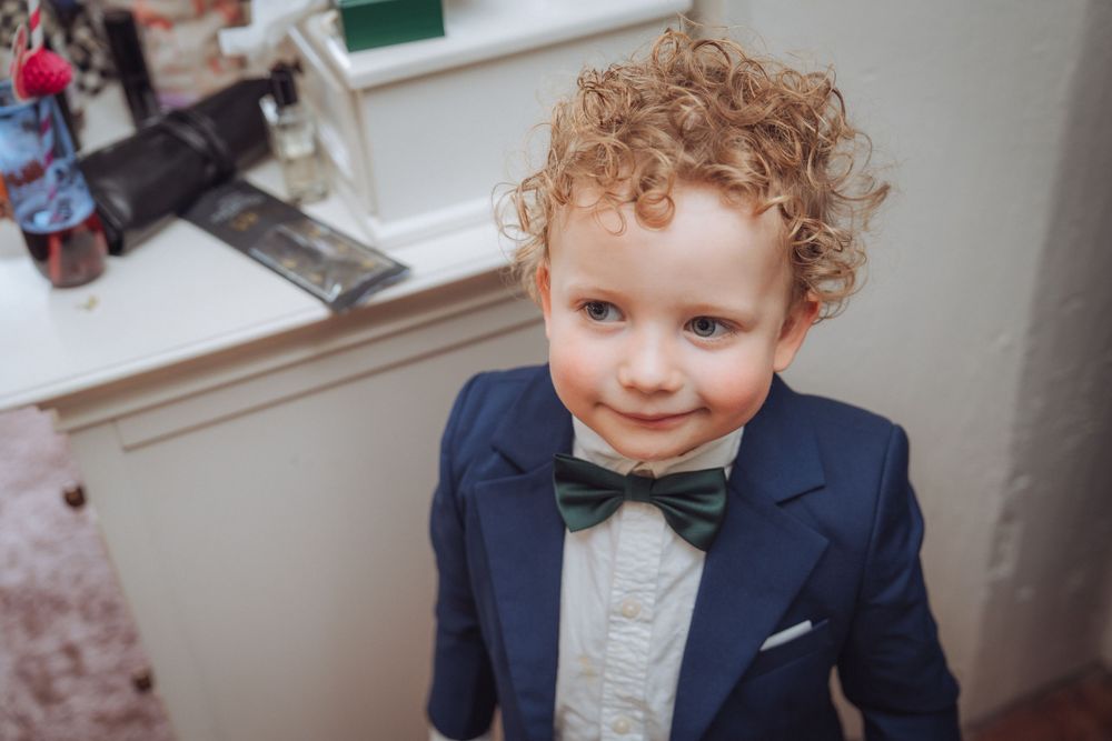 Adorable young boy with curly hair wearing a navy suit and green bow tie, smiling at a formal event.