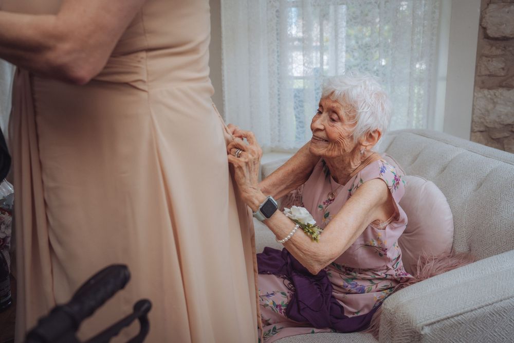 Elderly woman sitting on sofa touching bridesmaid dress at wedding preparation.