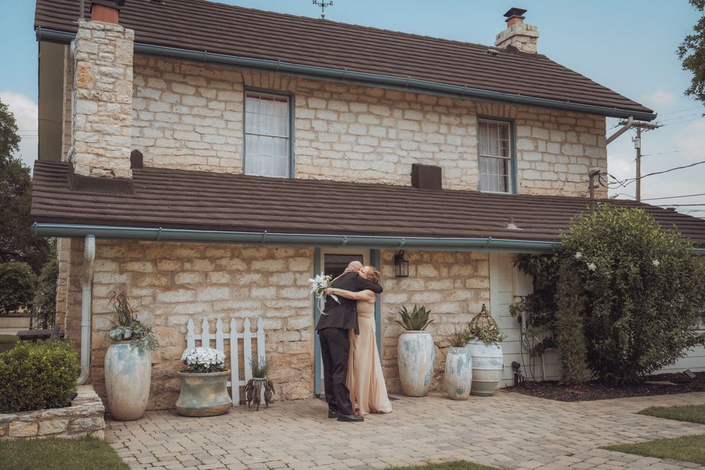 Couple embracing in front of a charming stone house with lush greenery, captured during a romantic outdoor wedding photoshoot.
