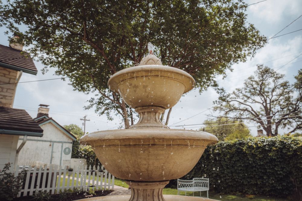 Two-tiered stone garden fountain with water flowing, surrounded by lush trees and hedges near a white fence.