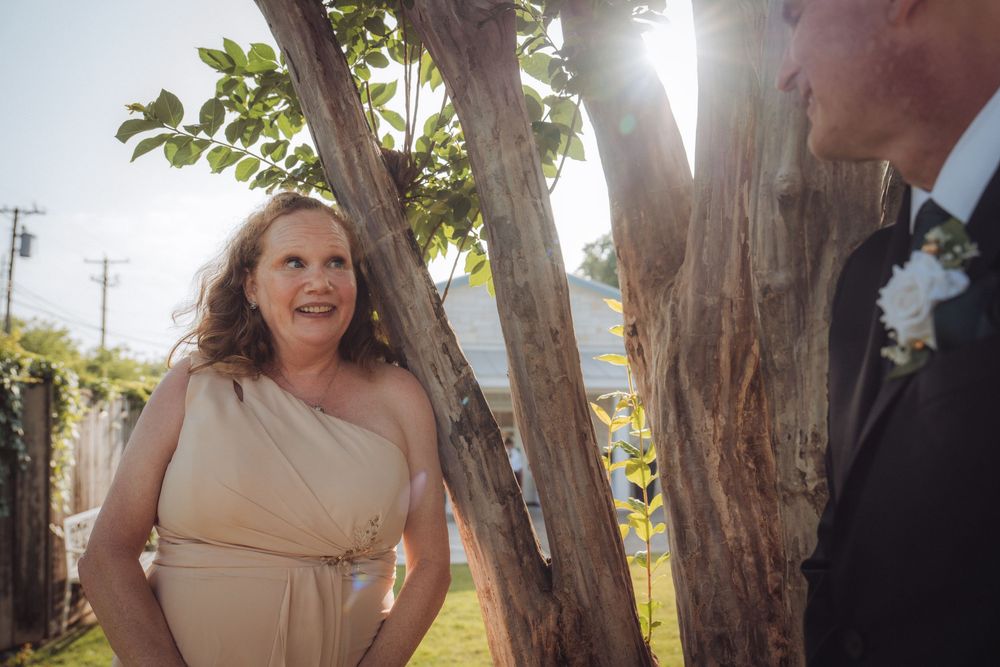 Woman in beige one-shoulder dress smiling at groom outdoors at sunny wedding ceremony near rustic tree.