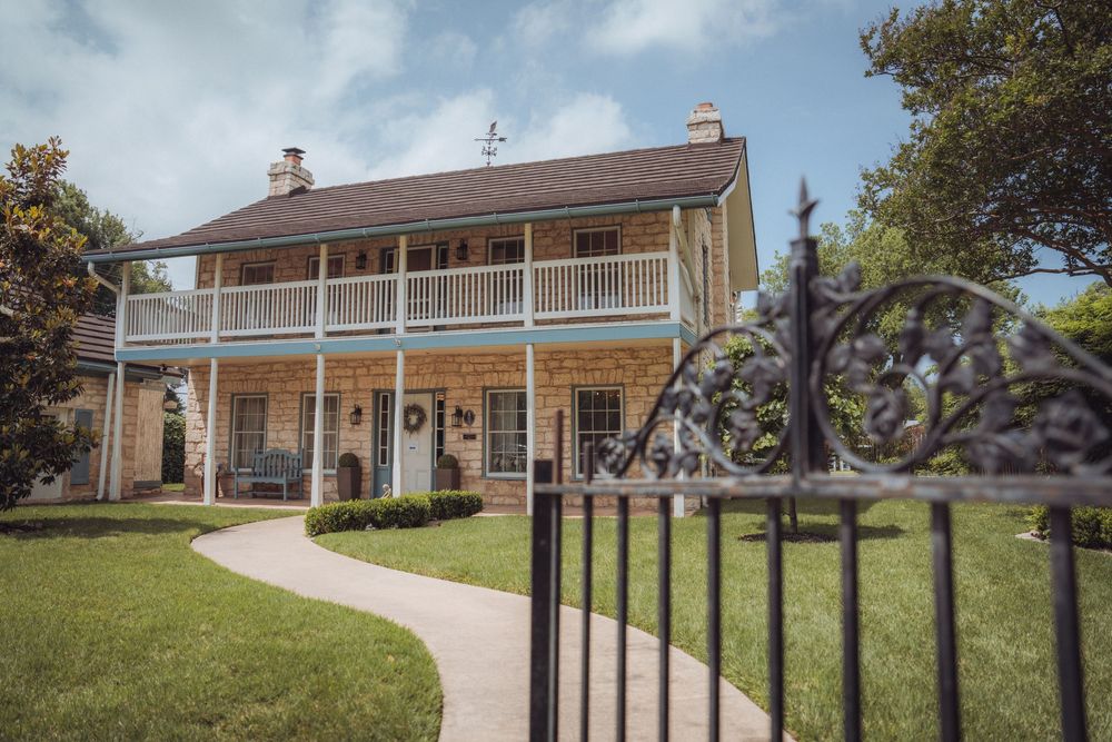 Historic two-story Southern plantation home with wrap-around balcony, brick facade, and ornate iron gate on lush green lawn.