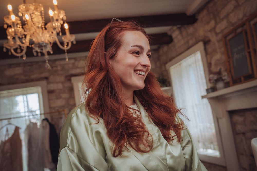 Smiling redhead woman in a sage green satin robe inside a rustic stone room with chandelier.