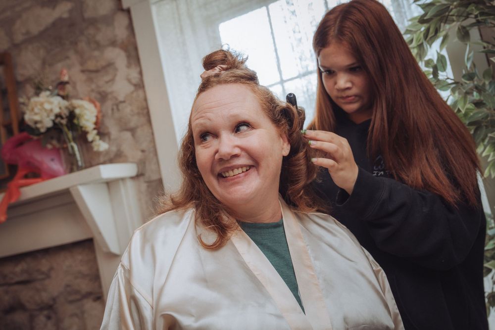 A woman in a satin robe smiles as a young girl with red hair styles her hair indoors.