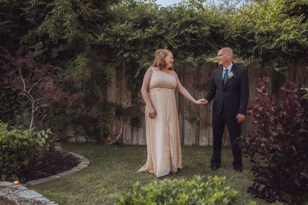 Couple in formal attire holding hands in a lush garden, woman in gold gown and man in dark suit with boutonniere.