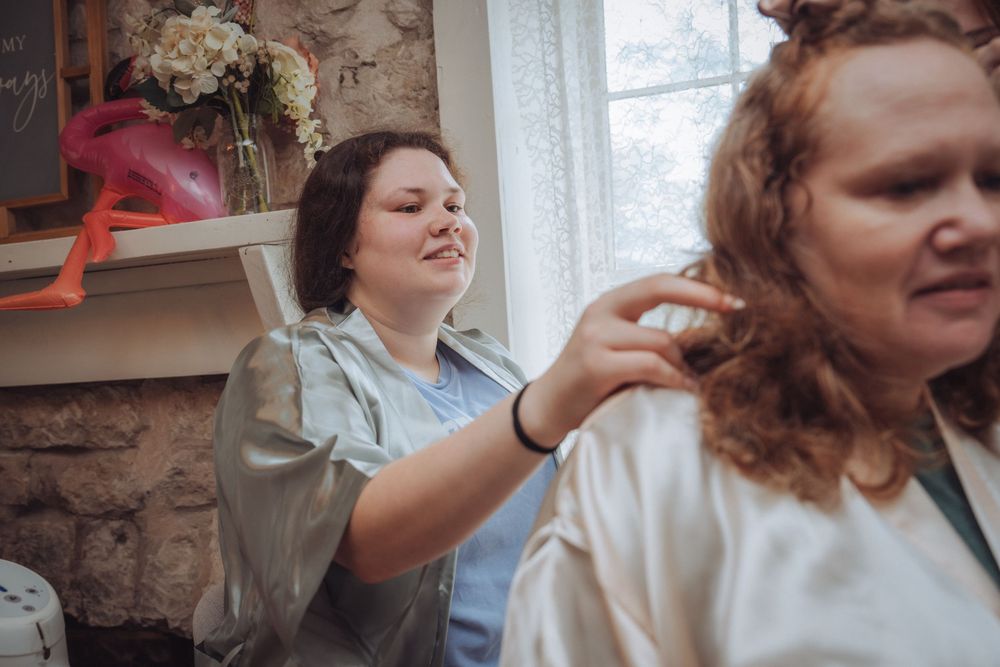 A hairstylist in a satin robe styles a woman's curly red hair near a window in a cozy stone-walled bridal suite.