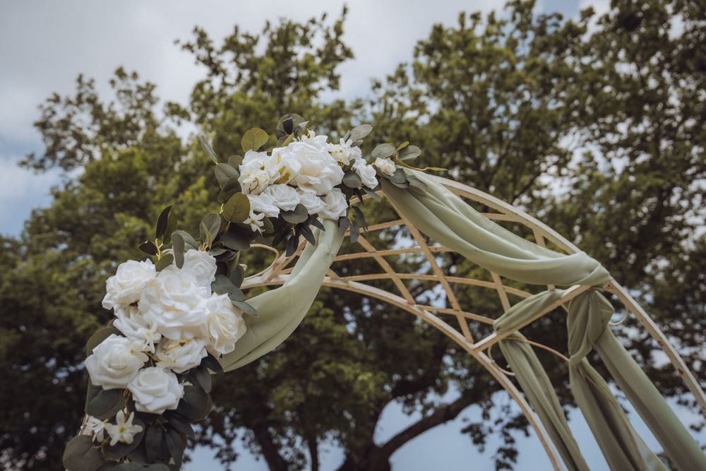 Elegant wedding arch adorned with white flowers and sage green fabric draped over woven branches outdoors.