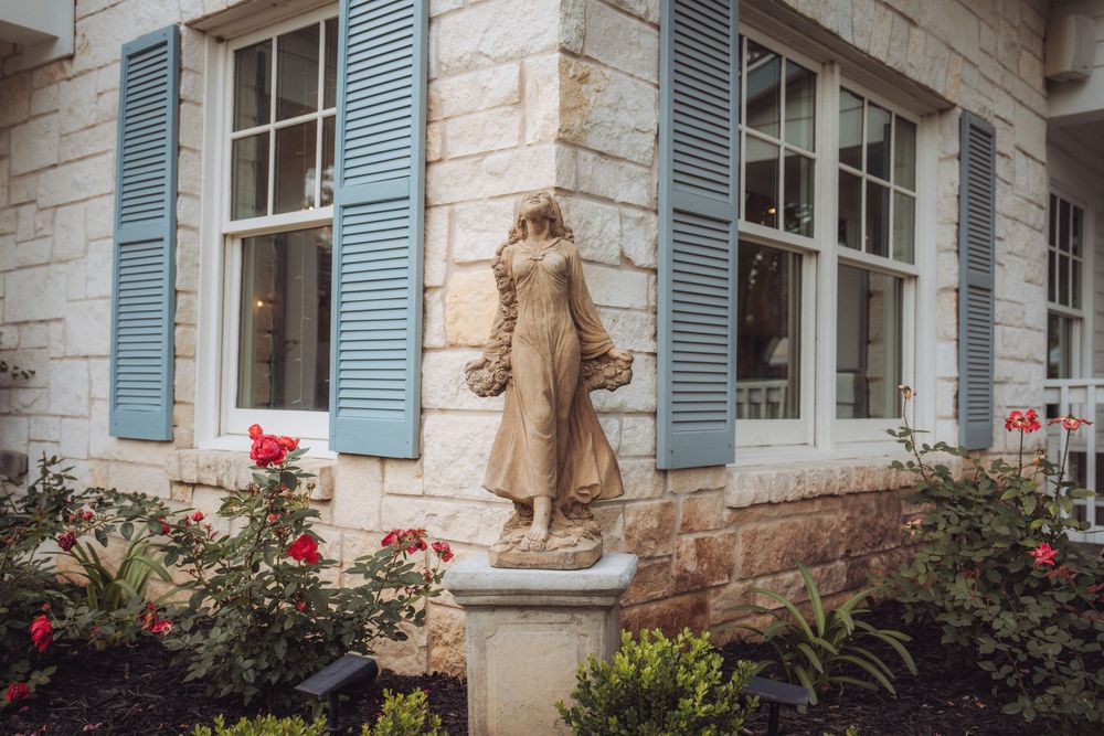 Stone statue of a robed woman on a pedestal, framed by red roses and blue shuttered windows of a limestone building.