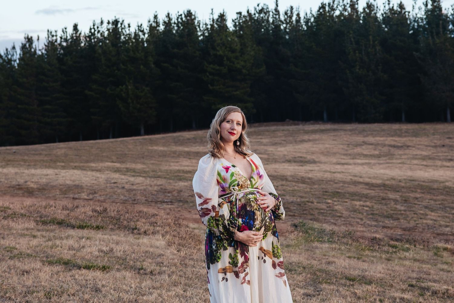 Canberra maternity in a white dress holds a vibrant floral bouquet while standing in a brown field with pine trees in the background.