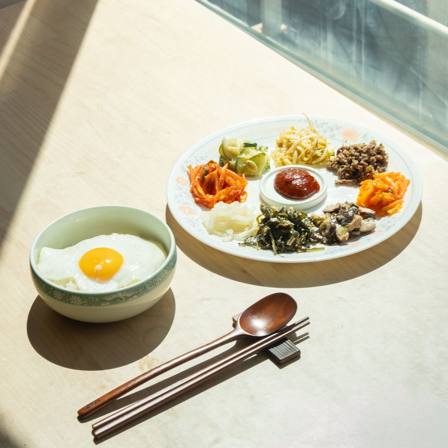 Korean bibimbap ingredients served on a white plate with sunny side up egg in green bowl, chopsticks and spoon on table.