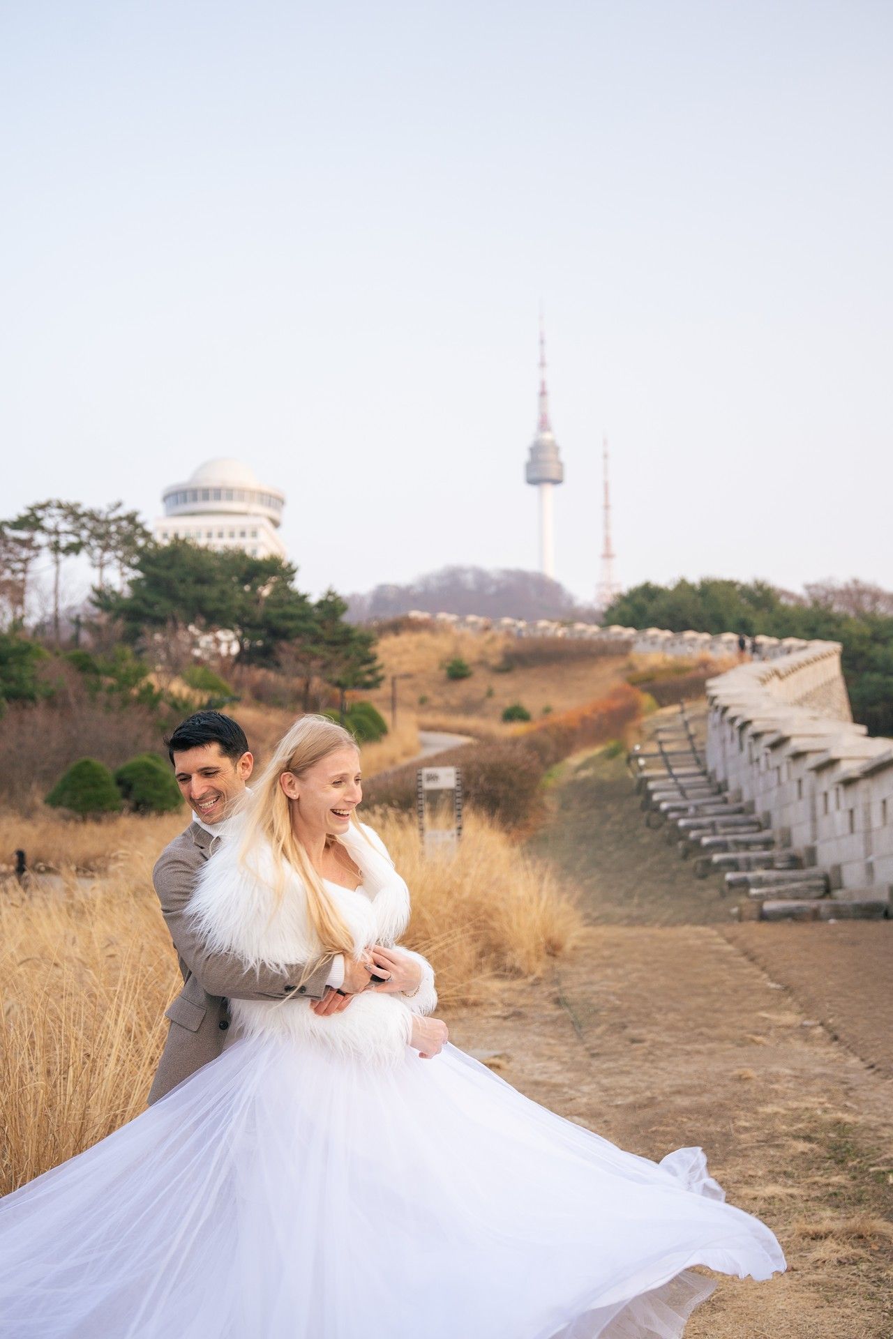 A couple in wedding attire dances together near Seoul Tower and ancient fortress walls in South Korea.