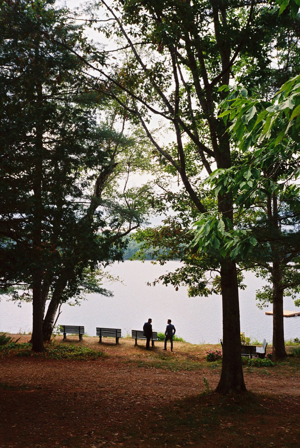 The groom and his best man talk intimately together before the ceremony under the trees by the lake during their Readville wedding in Maine.