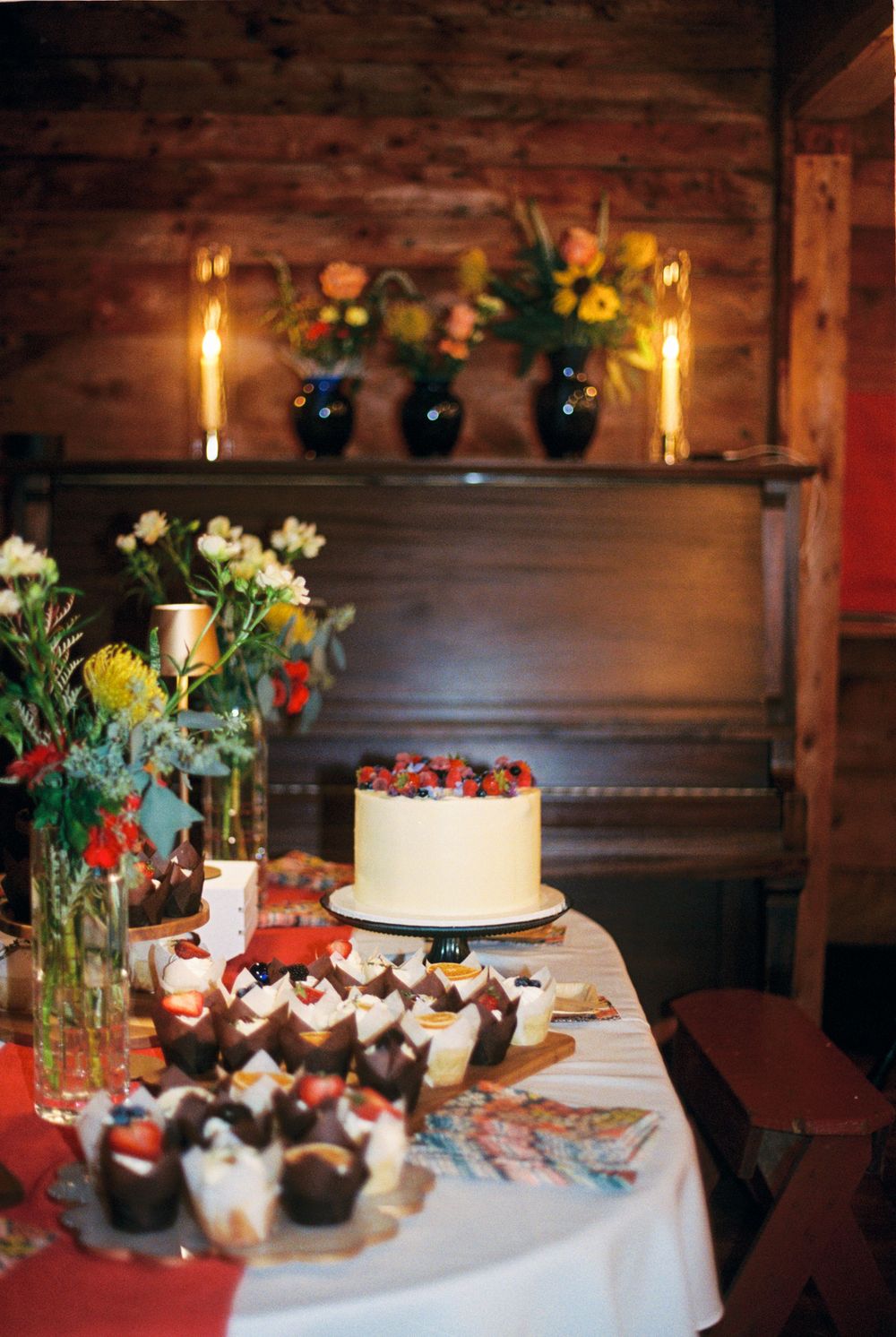 The wedding cake is centered and surrounded by delicious cupcakes, flowers, and in the background an upright piano with vases and candles.