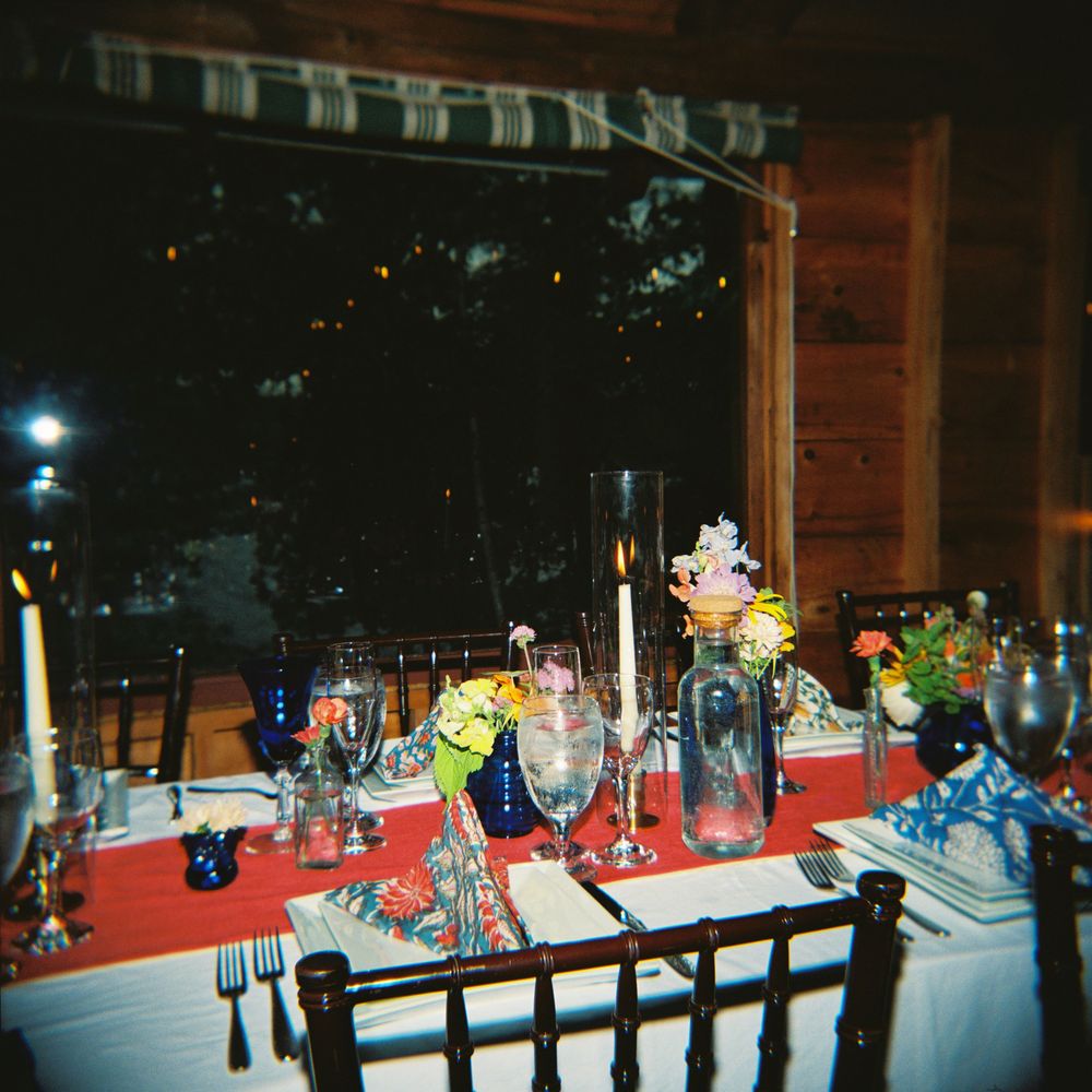 120mm color flash photograph of the bride and groom table at a camp wedding with flowers and red accents an blue vintage glassware in Maine.