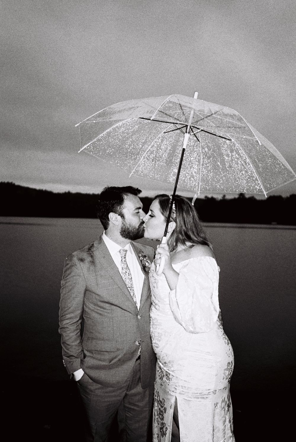 Black and white 35mm film portrait of the bride and groom kissing under a clear umbrella during a very rainy wedding in Readville, Maine.