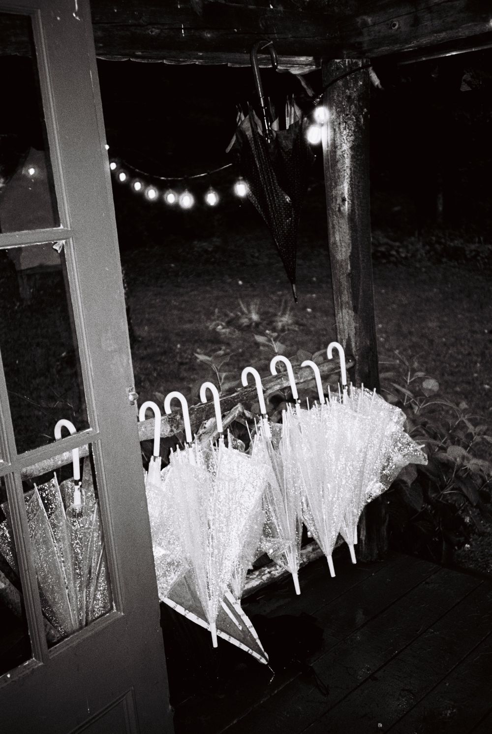 A row of umbrellas rests on the banister outside a wedding reception due to heavy rains while a string of lights shine on black & white film