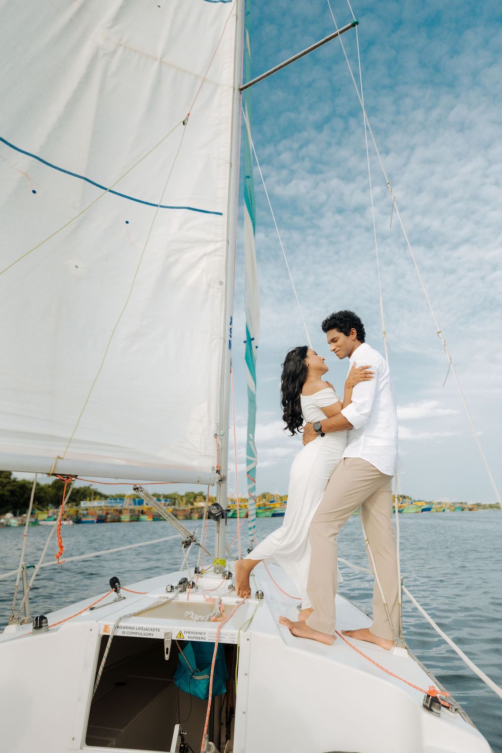 A couple shares intimate moments on a sailboat deck with white sails and blue skies in the background.