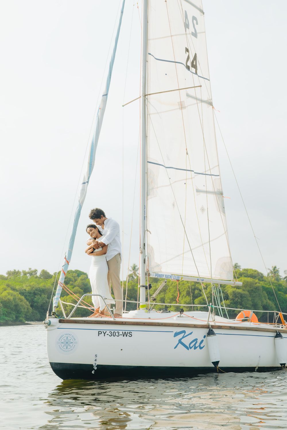 White sailboats float on tranquil waters near a green shoreline during a couple's photoshoot.