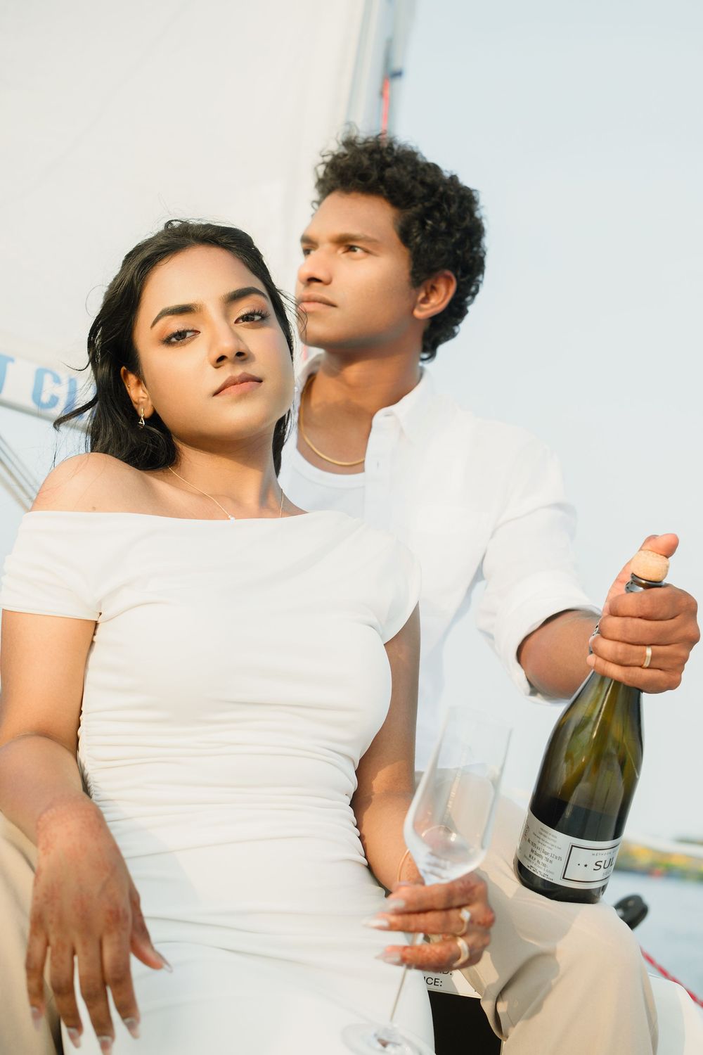 A couple in white outfits shares an intimate moment while holding champagne glasses outdoors.