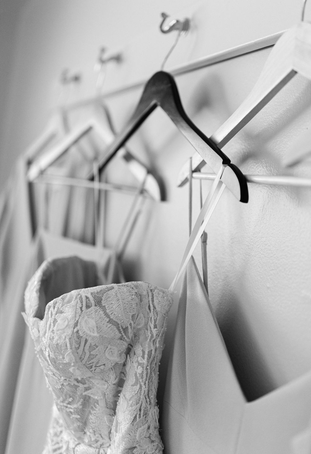 A delicate lace wedding dress hangs on a black hanger against a white wall with other hangers visible in the background.