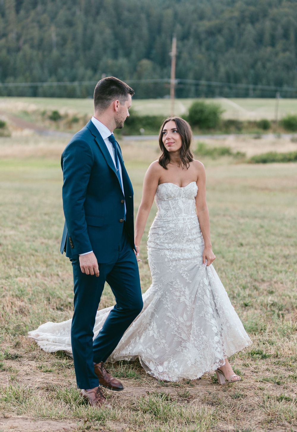 A couple in wedding attire poses playfully in a grassy field with mountains in the background.