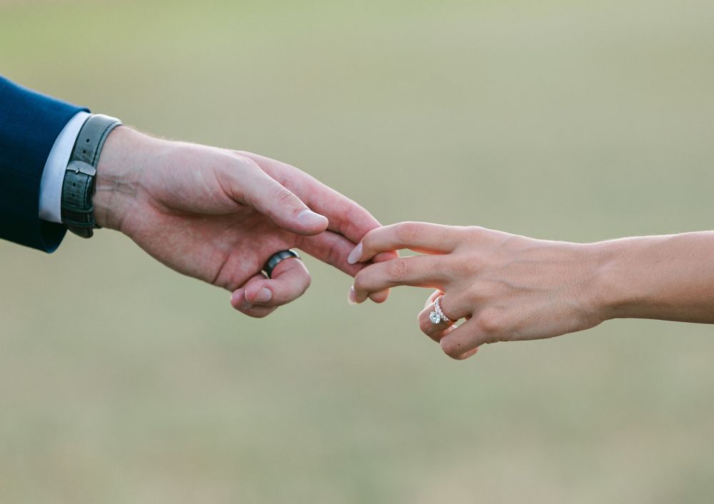 Two hands reaching towards each other with wedding rings in a romantic outdoor setting.