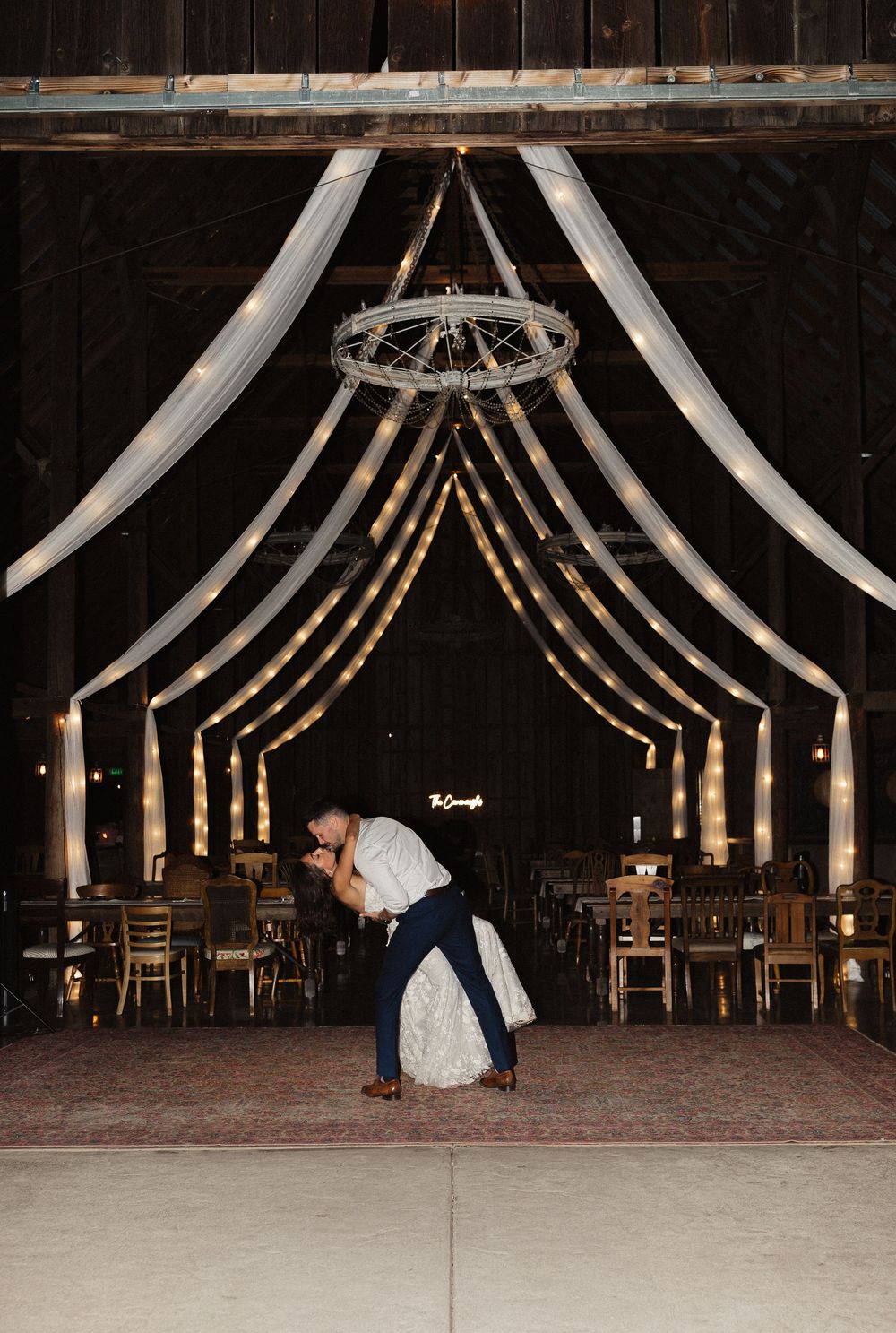Wedding couple dances in a rustic barn venue decorated with elegant white fabric draping and chandeliers.