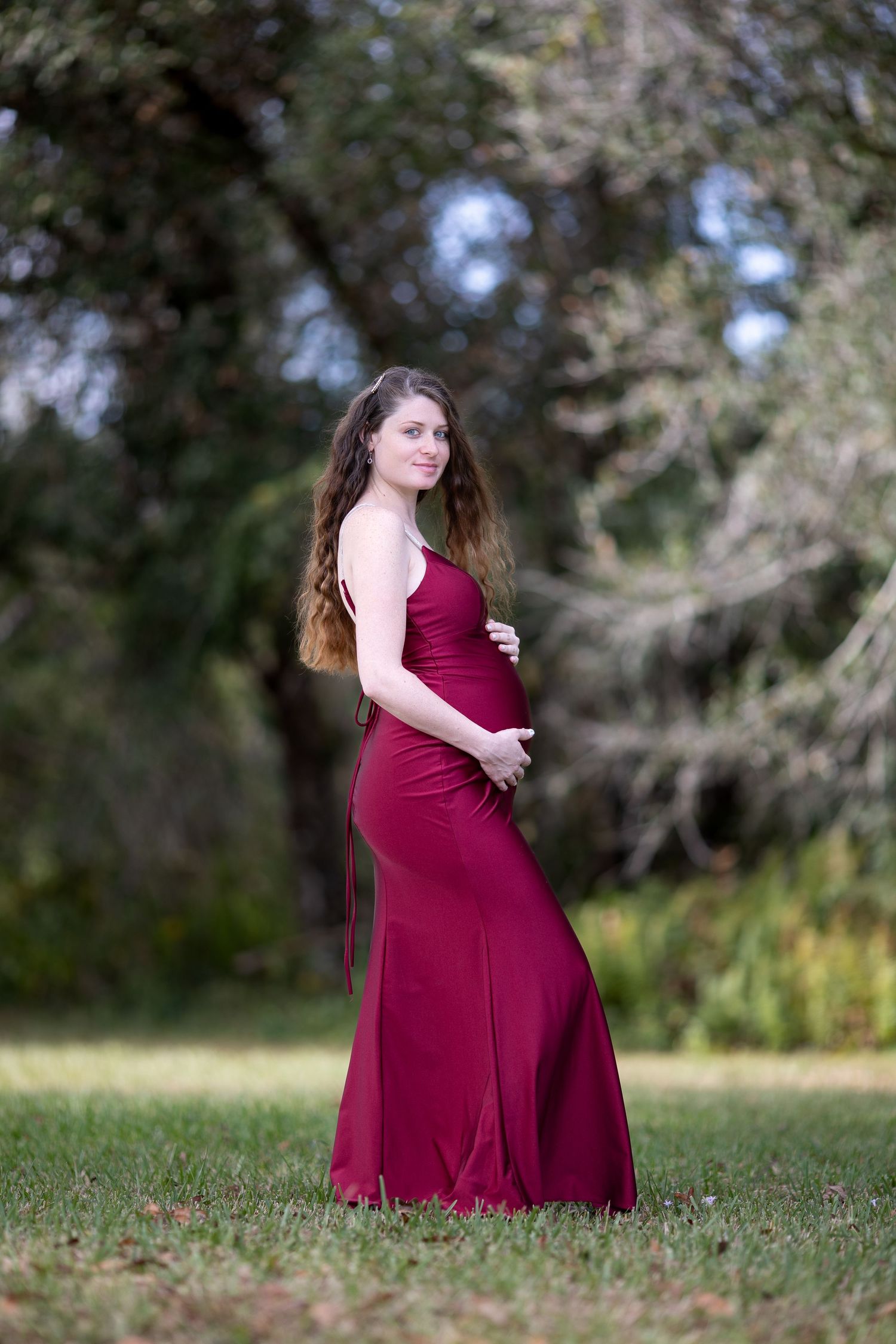 A woman in a burgundy dress poses gracefully on grass during an outdoor maternity photo session.