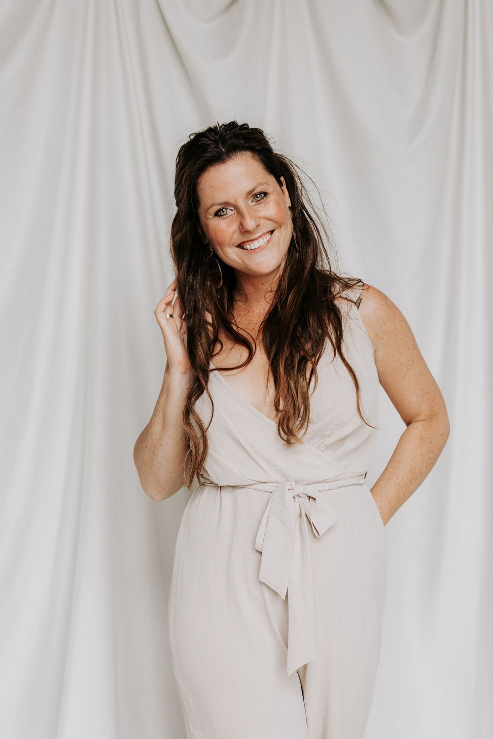 Smiling brunette woman in beige jumpsuit posing against white fabric backdrop.