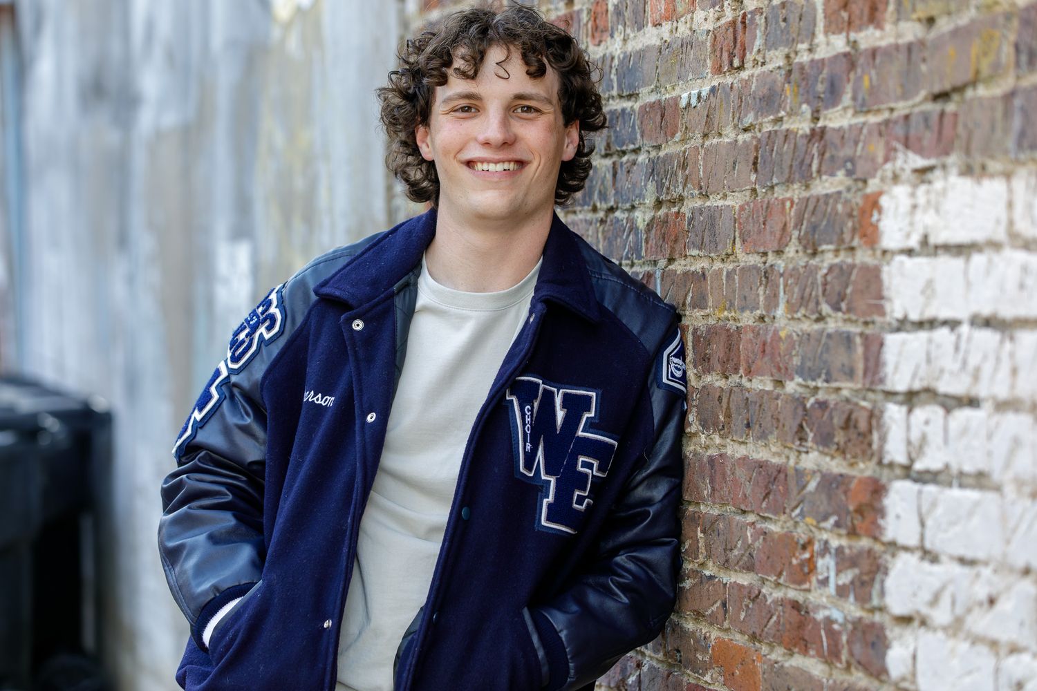 Young man smiling in a navy blue varsity jacket, leaning against a rustic brick wall in an alley.