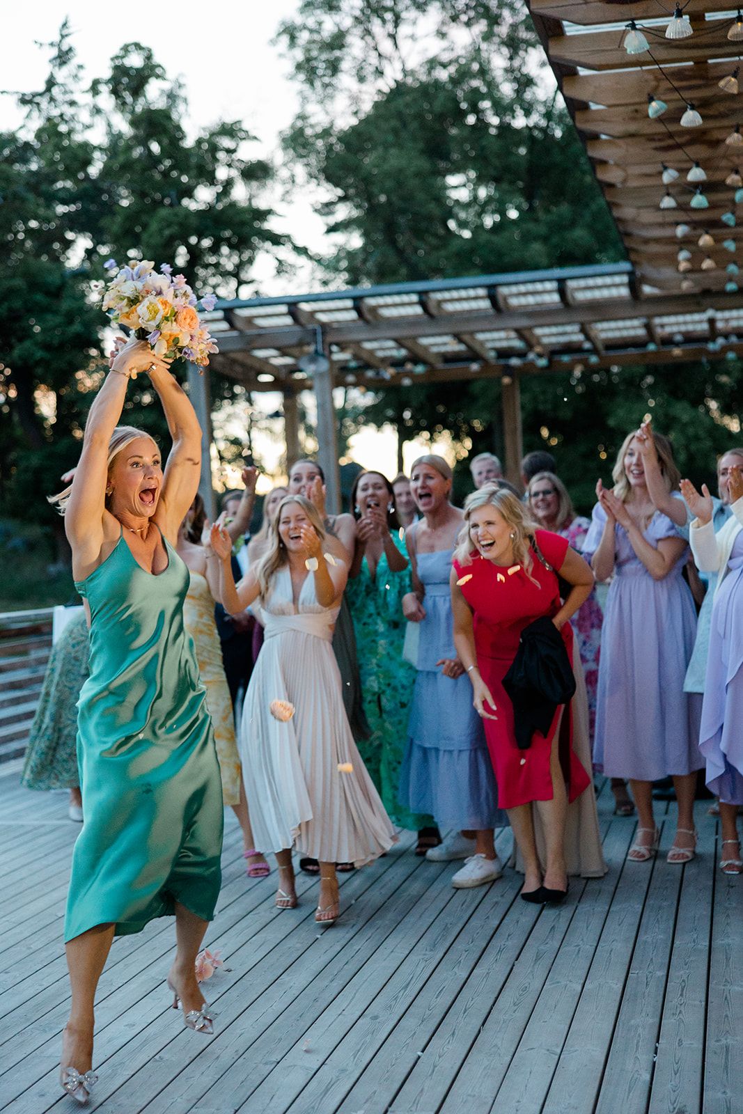 A group of wedding guests in colorful dresses gather on a wooden deck under string lights at an outdoor reception.