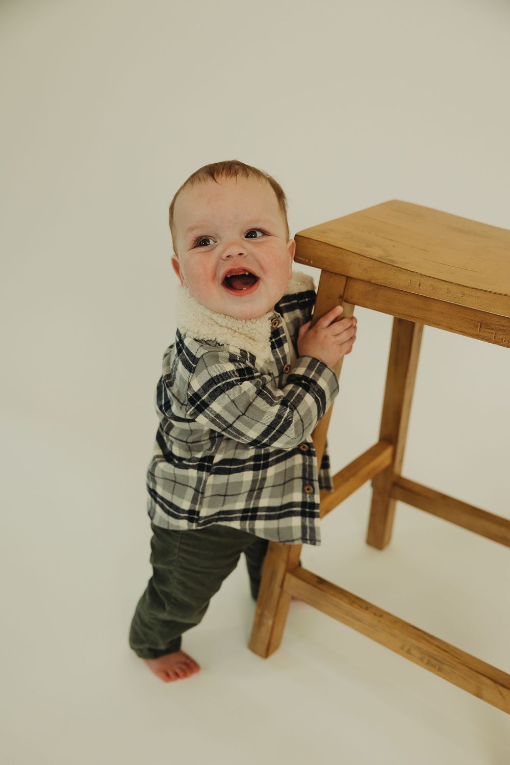 A baby in a plaid shirt and dark pants crawls and plays near a wooden bench in a light-colored room.