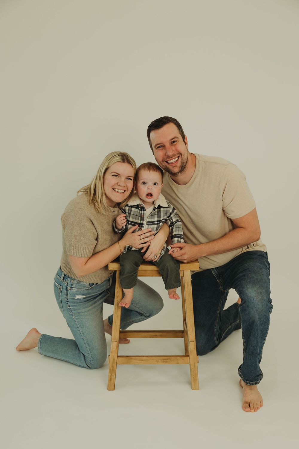 A family poses together on the floor in a minimalist studio setting with a small wooden stool.