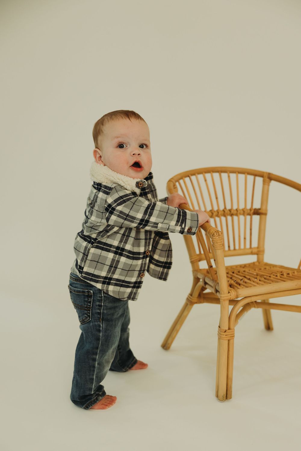 A baby in plaid shirt and jeans explores a wooden rattan bench while learning to stand.