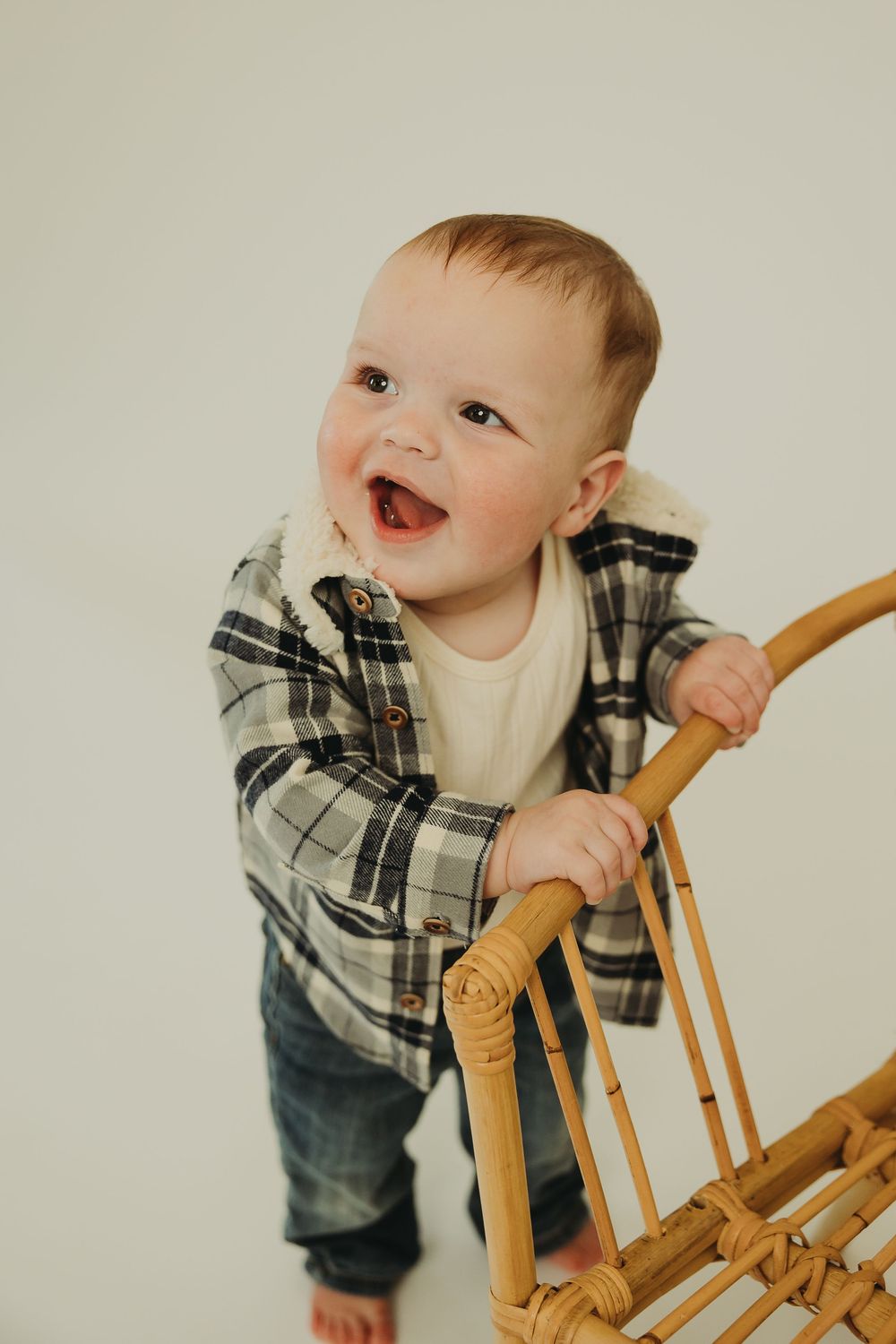 Baby in plaid shirt and jeans smiles while holding onto wooden chair in studio setting.