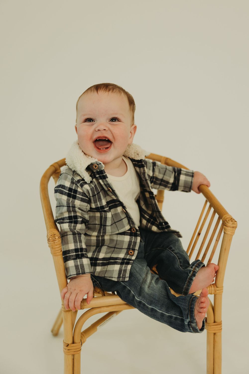 Baby wearing plaid shirt and jeans sits and laughs in wooden chair against beige background.