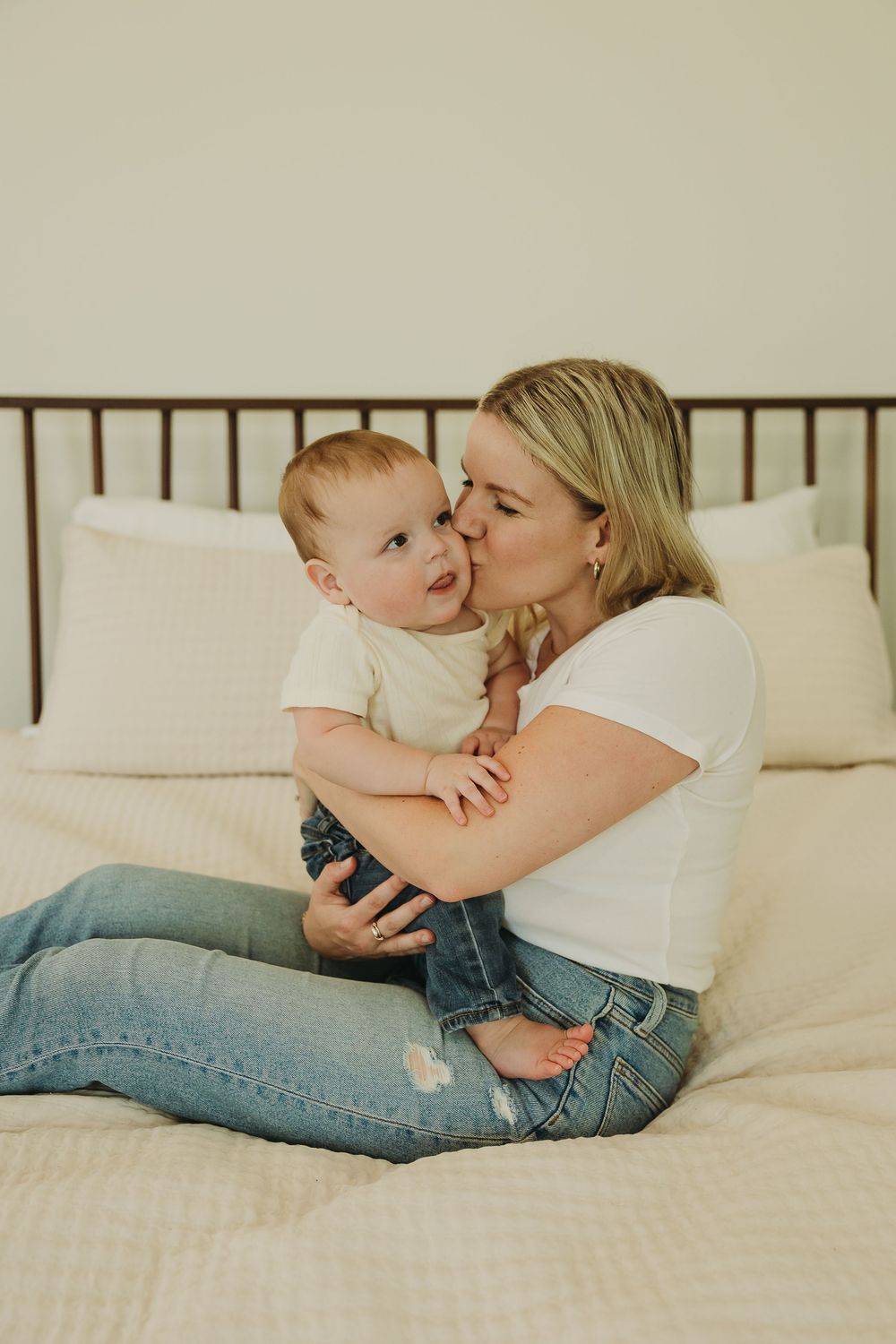 Parent and baby share tender moments while relaxing together on white bedding in casual denim attire.