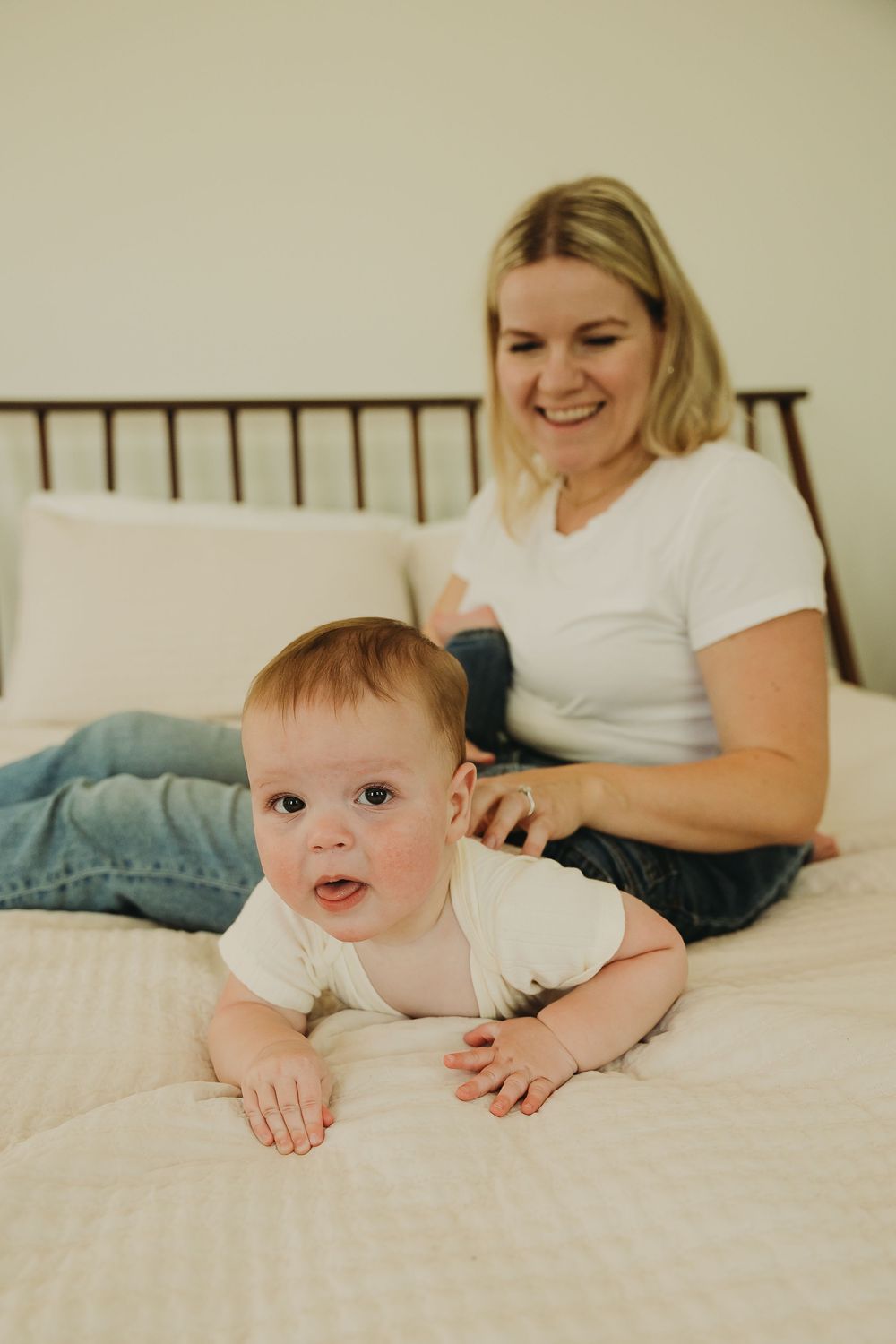 Baby in white onesie lies on white bedding while someone relaxes nearby in casual attire.