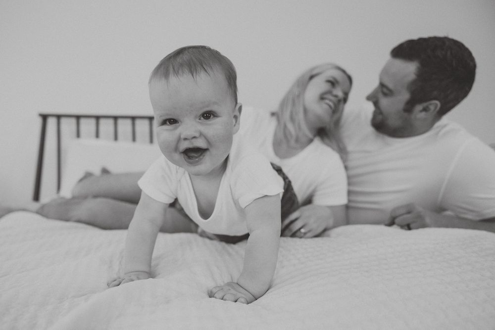 A happy baby crawls on a bed while parents relax in the background in black and white photography.