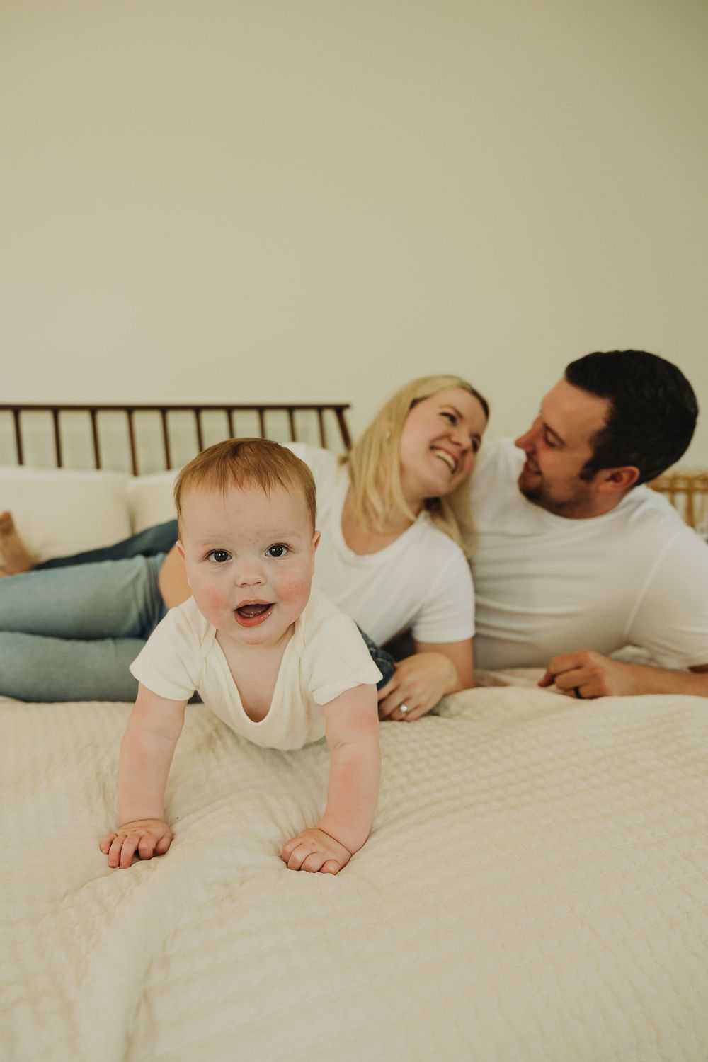 A happy family relaxes together on a white bed, with a joyful baby crawling on the mattress.
