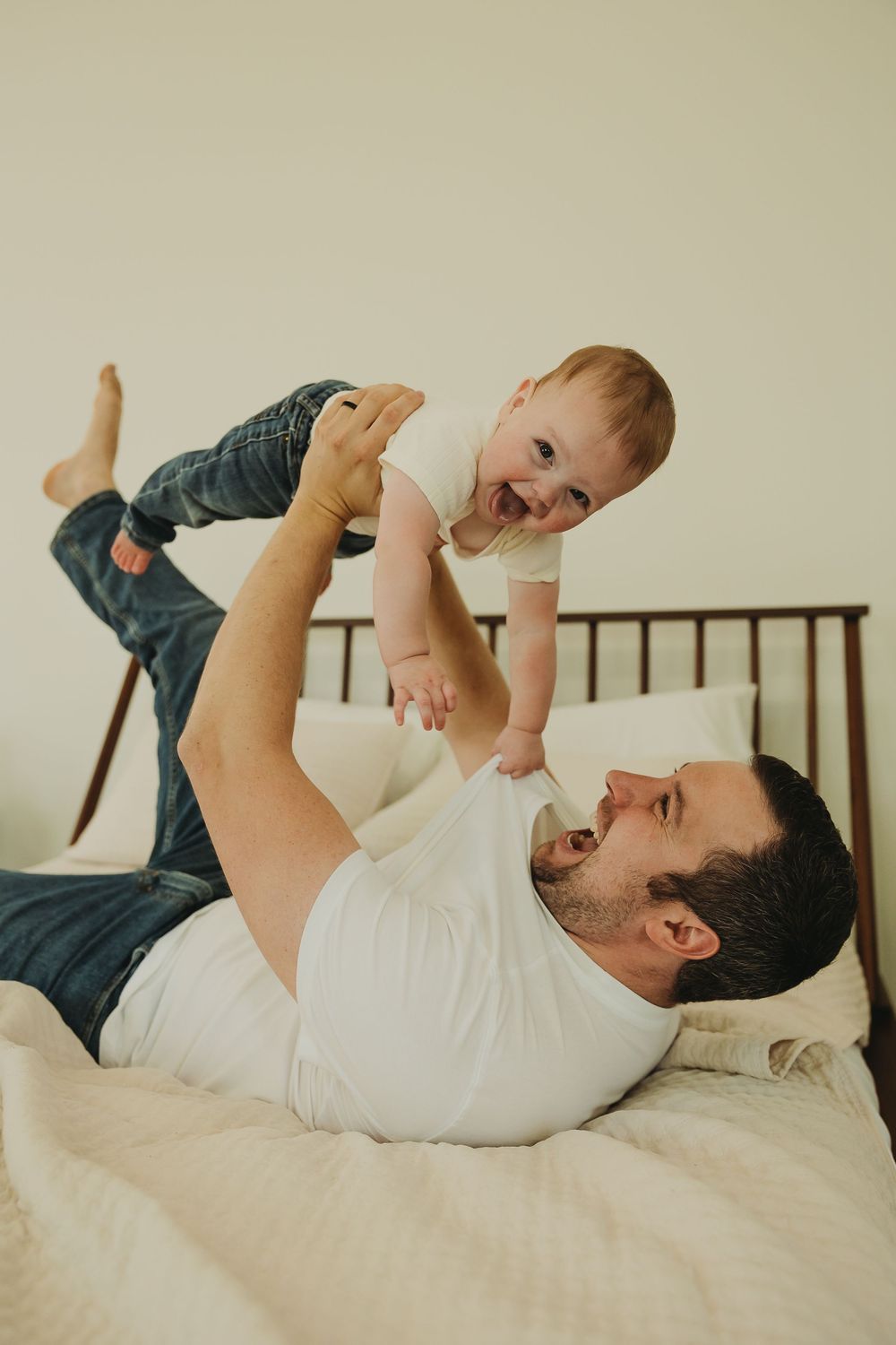 A parent and baby share a joyful moment playing together while lying on a white bed.