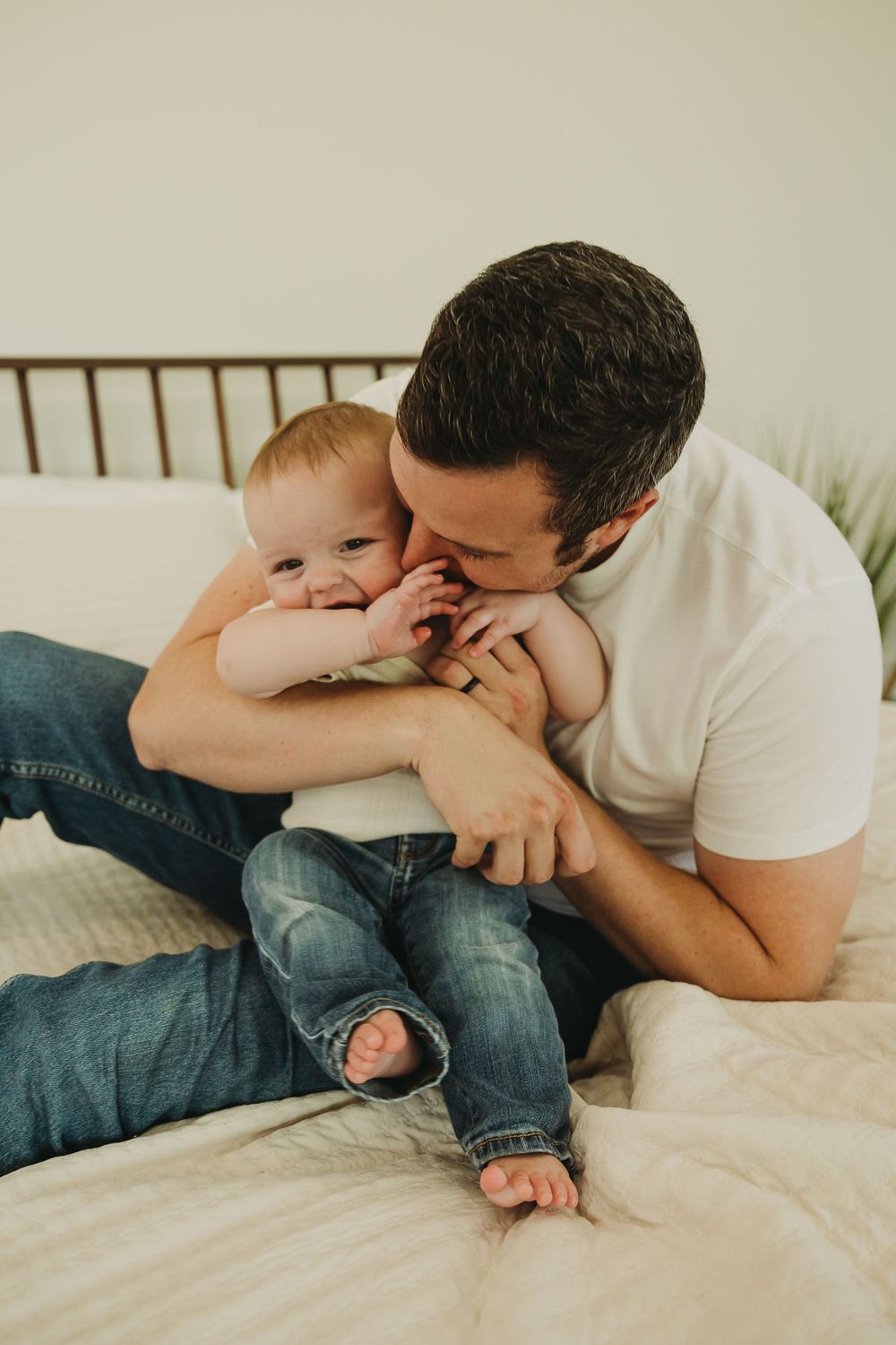 Parent and baby share tender playful moments while lying together on white bedding.