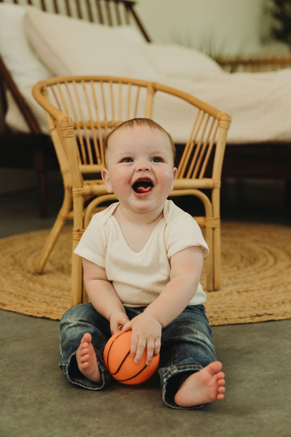 Laughing baby sits on round jute rug with small basketball while wearing white shirt and jeans.