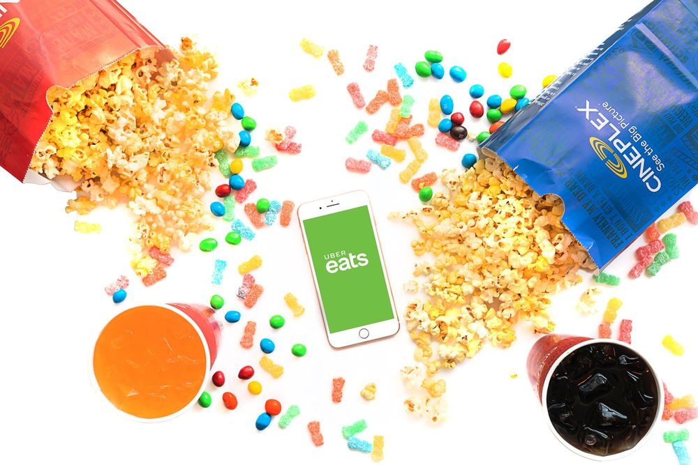 Colorful candy and popcorn scattered on white background. A phone with Uber Eats shown on the screen and popcord bags with Cineplex branding