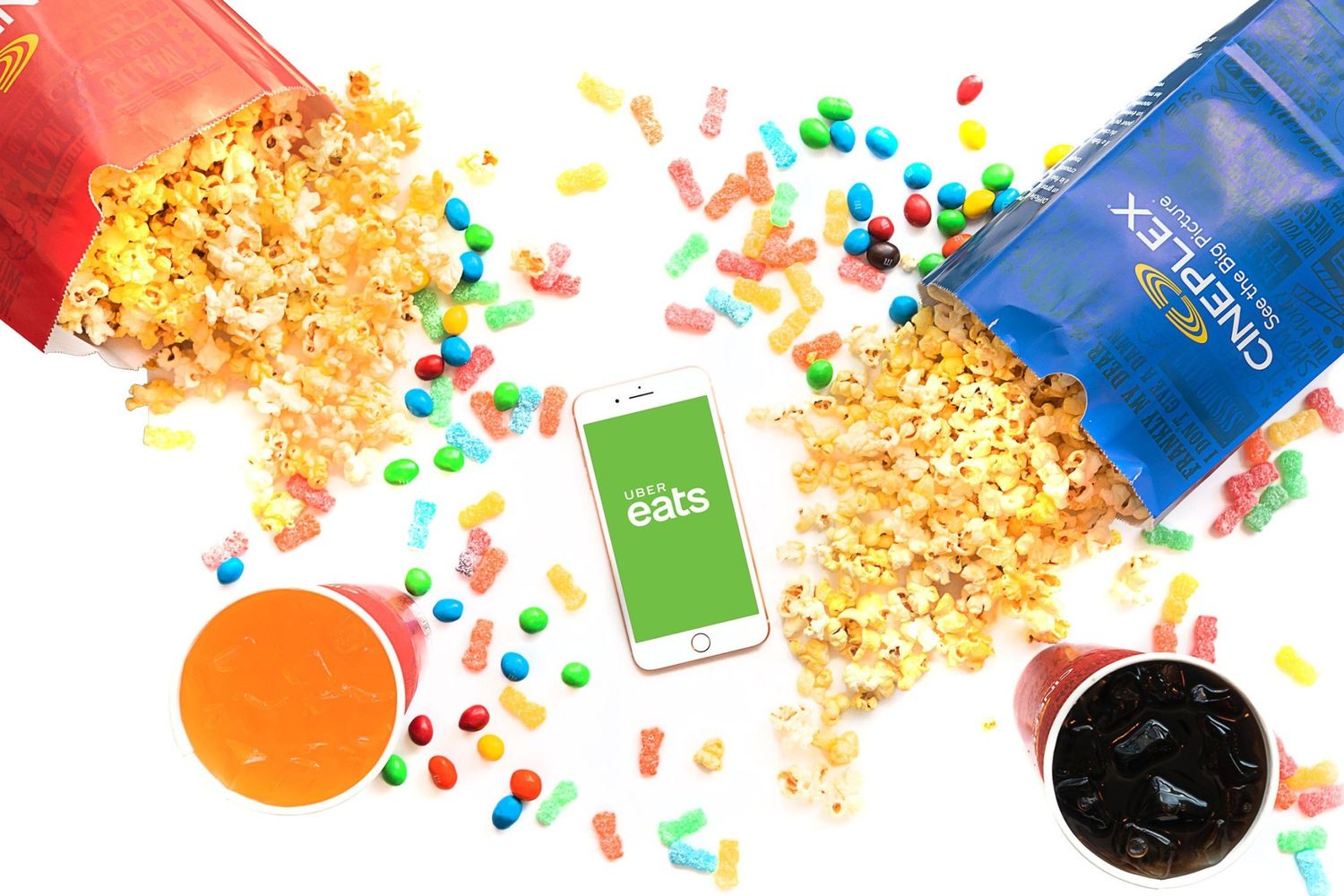 Colorful candy and popcorn scattered on white background. A phone with Uber Eats shown on the screen and popcord bags with Cineplex branding
