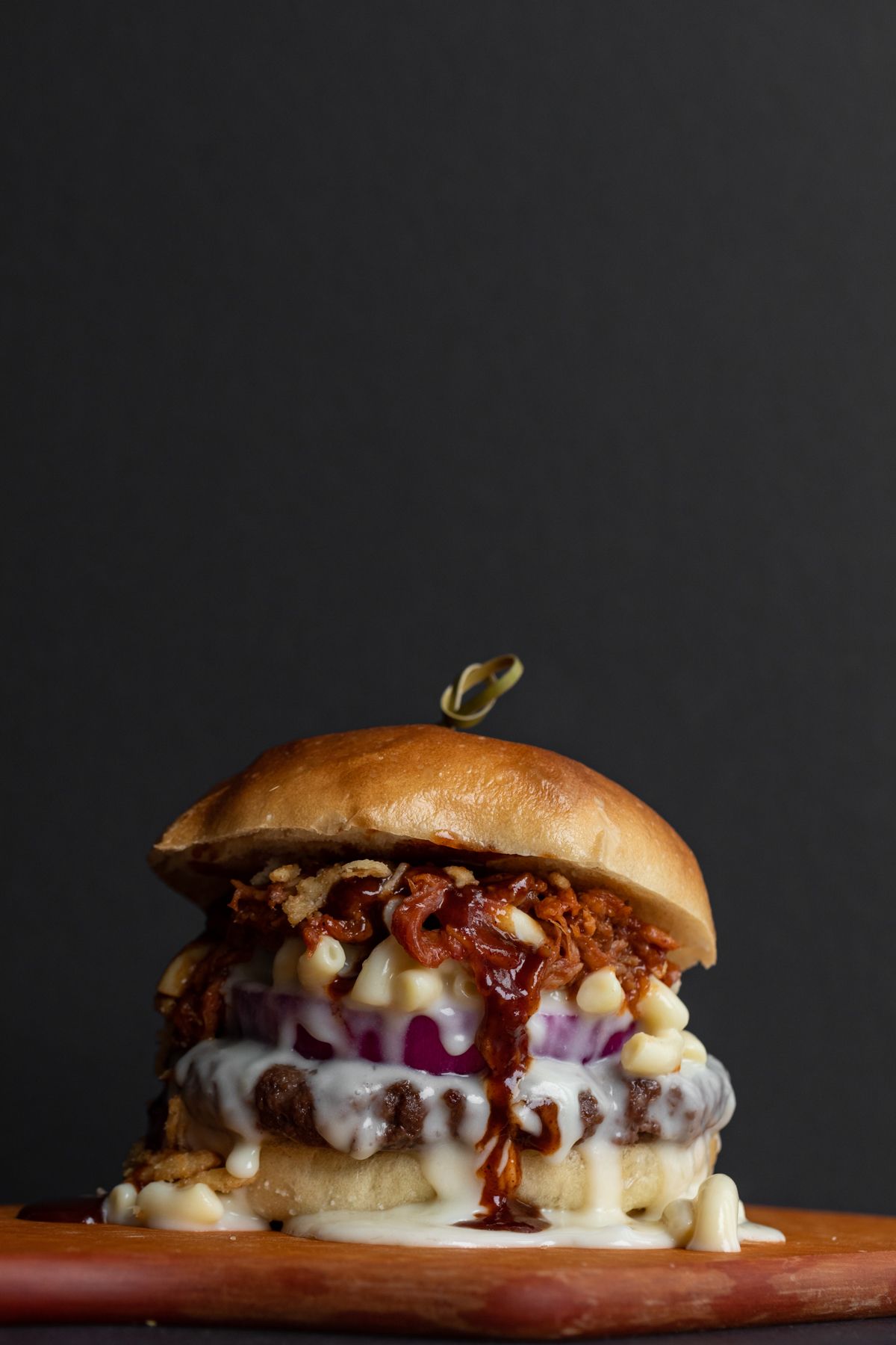 Gourmet burger with melted cheese and sauce on wooden board against dark background.